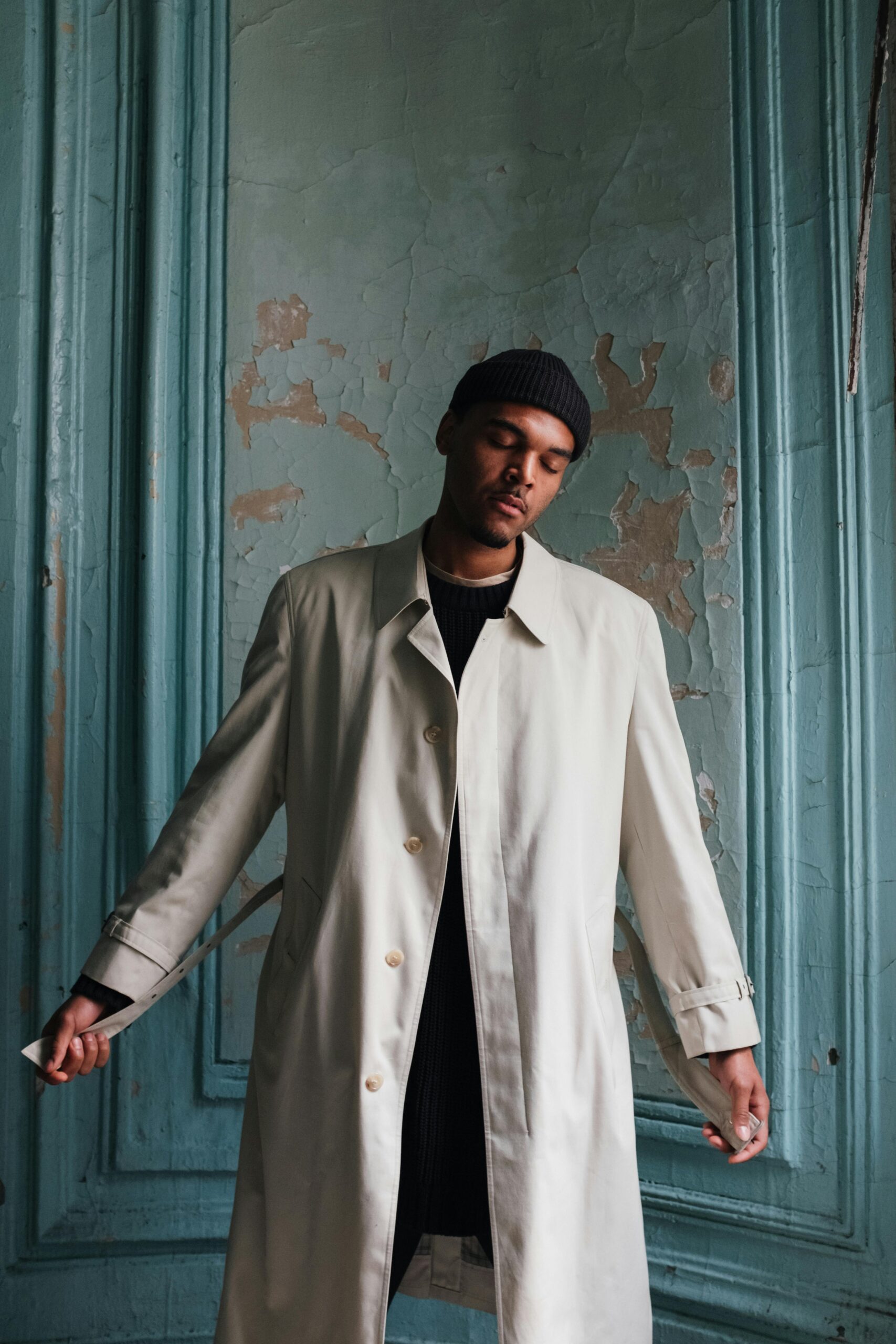 Fashion portrait of a man in a beige coat standing indoors against a rustic, textured wall.